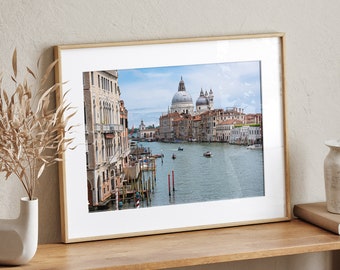 May include: A framed photograph of a canal in Venice, Italy. The photo shows a view of the Grand Canal with the Basilica di San Giorgio Maggiore in the background. The photo is framed in a light wood frame.