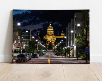 May include: A night view of a city street with a large building in the distance. The street is lined with trees and streetlights, and there are cars parked on the side of the road. The building is illuminated by lights, and there is a crescent moon in the sky.
