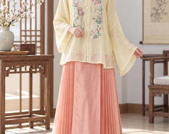 May include: A pale yellow long-sleeved blouse with a square neckline and floral embroidery on the front, paired with a floor-length pleated peach skirt featuring a white floral border at the hem. The blouse has a button-down front with small pearl buttons.