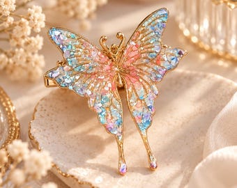 May include: A gold-toned butterfly hair clip featuring iridescent, multi-colored flake inlays in shades of pink, blue, and purple across the wings. The butterfly has a delicate metal frame with two antennae and elongated lower wing tips.