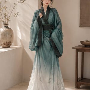 Peut inclure: Une robe longue au sol présentant un dégradé de couleurs bleu sarcelle et blanc avec des manches larges et fluides. Le vêtement comprend un col à motifs vert foncé et une large ceinture assortie, avec des motifs de feuilles de bambou.