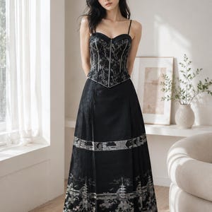 Pode incluir: Um vestido de noite preto com um corpete estilo espartilho e uma saia fluida. O corpete apresenta bordados prateados intrincados, enquanto a saia mostra um design detalhado de paisagem de inspiração asiática. O vestido tem finas alças espaguete.
