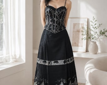 May include: A black evening gown with a corset-style bodice and a flowing skirt. The bodice features intricate silver embroidery, while the skirt showcases a detailed Asian-inspired landscape design. The dress has thin spaghetti straps.