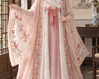 May include: A floor-length, multi-layered dress in shades of dusty pink and cream, featuring intricate floral embroidery on the sheer outer robe and bodice. The garment includes wide, flowing sleeves, a tied waist sash, and a pleated skirt overlay.