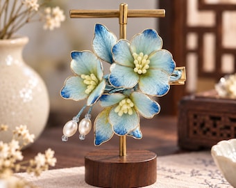 May include: A hair clip featuring three blue and white gradient flowers with yellow centers, accented by two dangling white oval pearls on thin blue threads. The floral hair accessory is mounted on a gold-toned metal T-shaped display stand with a round wooden base.