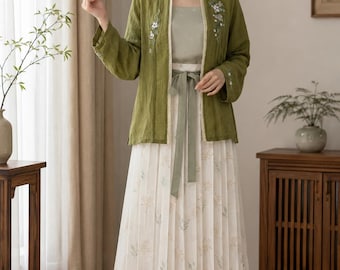 May include: An olive green open-front jacket with floral embroidery, worn over a sage green camisole and a long, white pleated skirt featuring a delicate botanical print. The skirt is cinched at the waist with a matching sage green fabric tie.