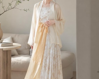 May include: A cream-colored, layered dress with a floral pattern and pleated skirt. The outfit includes a matching sheer jacket and a gold-colored sash tied at the waist. The model is wearing cream-colored flats.