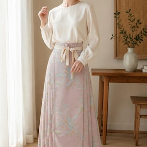 May include: A long-sleeved cream-colored blouse with a round neckline paired with a high-waisted, floor-length dusty pink skirt featuring a subtle botanical print in light blue and green, finished with a matching fabric tie belt at the waist.