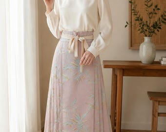May include: A long-sleeved cream-colored blouse with a round neckline paired with a high-waisted, floor-length dusty pink skirt featuring a subtle botanical print in light blue and green, finished with a matching fabric tie belt at the waist.