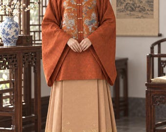 May include: A rust-colored long-sleeved top with a square neckline and a central panel featuring a colorful bird and cloud pattern, secured with small toggles. It is paired with a floor-length tan pleated skirt decorated with gold crane embroidery.