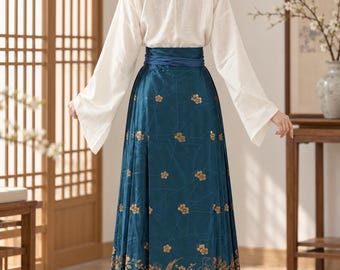 May include: A two-piece outfit consisting of a long-sleeved white top with a high collar and a floor-length teal skirt. The skirt features a dark blue waistband and a gold-colored floral and bird pattern along the hem and throughout the fabric.