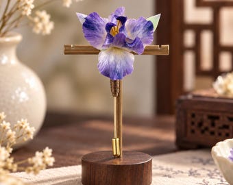 May include: A lapel pin featuring a realistic purple and white iris flower attached to a thin gold-toned metal stem. The pin is displayed upright on a T-shaped gold-toned metal stand, which is mounted on a circular dark brown wooden base.