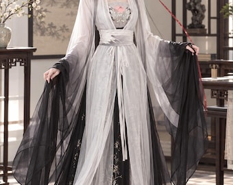 May include: A floor-length dress featuring a black base with intricate floral embroidery, layered with a sheer white and black gradient overskirt. The garment includes long, flowing sleeves, a wide waist sash, and a bodice with floral detailing.