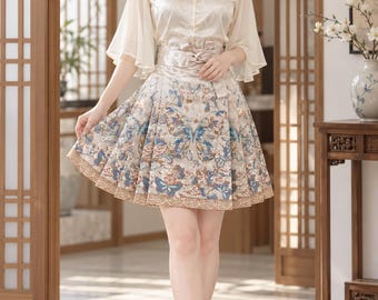 May include: A two-piece outfit consisting of a cream-colored satin blouse with a mandarin collar, tassel accents, and sheer flared sleeves, paired with a high-waisted, pleated skirt featuring a detailed blue and brown butterfly and floral pattern.