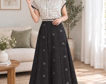 May include: A two-piece outfit consisting of a short-sleeved cream blouse with a black diamond pattern and black trim, paired with a high-waisted, floor-length charcoal gray skirt featuring a subtle embroidered butterfly motif and a thin white belt.