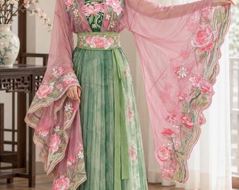 May include: A floor-length dress featuring a green pleated skirt and a matching green bodice with pink floral embroidery. The garment includes a sheer, flowing pink outer layer with wide sleeves and intricate pink and green floral embroidery throughout.