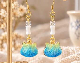 May include: A pair of gold-toned hook earrings featuring a cat-shaped charm with a blue and yellow gradient finish. Each earring includes a clear spherical bead and a white cylindrical bead suspended above the cat charm on a gold metal wire.