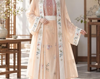 May include: A peach-colored ensemble featuring a long, sheer outer robe with white embroidered trim and wide sleeves, worn over a matching pleated skirt and a sleeveless bodice with a square neckline, white buttons, and lace detailing.