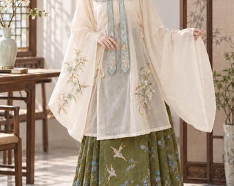 May include: A traditional-style ensemble featuring a cream-colored, long-sleeved sheer top with a high collar, light blue embroidered trim, and floral motifs, paired with a floor-length olive green skirt decorated with blue flowers and birds.