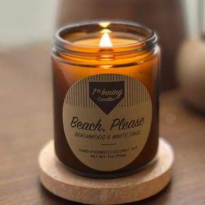 May include: A lit candle in a clear glass jar with a brown label that reads "Beach, Please". The candle sits on a round wooden coaster. The background includes a wooden vase and a glass vase with flowers.