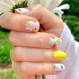 May include: A hand model wearing nail wraps with a floral and bee design. The nail wraps are white with yellow, red, and green accents. The hand is holding a white daisy.