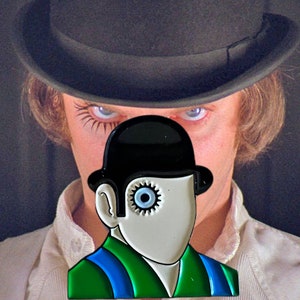 May include: A black and white enamel pin featuring a stylized face wearing a black bowler hat. The face has a gear for an eye and is set against a green and blue outfit. The background shows a person wearing a black hat.