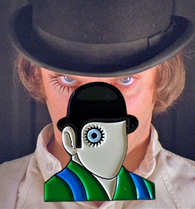 May include: A black and white enamel pin featuring a stylized face wearing a black bowler hat. The face has a gear for an eye and is set against a green and blue outfit. The background shows a person wearing a black hat.