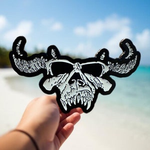 May include: Black and white skull with horns, resembling a bull's head, held in a hand against a beach background.
