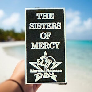 May include: A black rectangular sign with white text that reads "The Sisters of Mercy" and a white star with a face inside. The text "Merciful Release" is below the star.