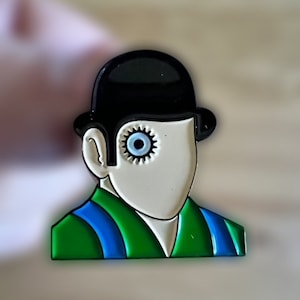 May include: A pin featuring a stylized face with a black bowler hat, a white face, and a gear-shaped eye. The figure wears a green jacket with blue stripes. The pin is a colorful, pop art style design.