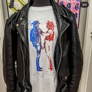 May include: A black leather motorcycle jacket worn over a light gray t-shirt. The shirt features a graphic of two figures in cowboy hats, one in blue and one in red, facing each other. The word DANCE is printed in light blue above the figures.