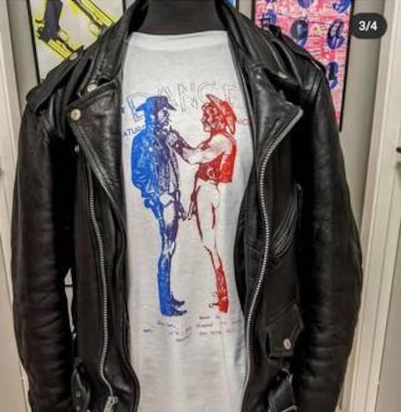 May include: A black leather motorcycle jacket worn over a light gray t-shirt. The shirt features a graphic of two figures in cowboy hats, one in blue and one in red, facing each other. The word DANCE is printed in light blue above the figures.