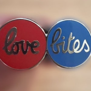 May include: A red and blue enamel pin with the words "love bites" in silver lettering.