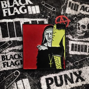 Puede incluir: Un pin cuadrado de esmalte que representa a una monja con hábito negro y cruz dorada, sobre un fondo rojo y amarillo. El pin está rodeado de imperdibles y un fondo con las palabras "BLACK FLAG" y "PUNX".
