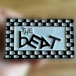 May include: Black and white enamel pin with a chequered border. The pin says "THE BEAT" in white letters.