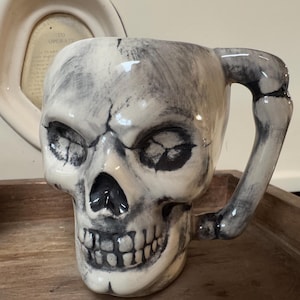 May include: A white ceramic skull mug with a black bone handle. The skull has black painted details, including eyes, nose, and teeth.