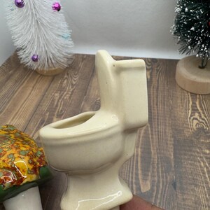May include: A white ceramic toilet shaped planter with a small opening at the top for planting. The planter is sitting on a wooden surface.