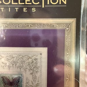 May include: A framed cross-stitch kit featuring a butterfly and floral design. The kit includes a purple background and a gold-colored frame. The text "COLLECTION" and "PETITES" is visible at the top.