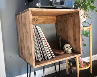 May include: A wooden record player stand with a black record player on top and a stack of vinyl records on the shelf. The stand has black hairpin legs and is sitting on a rug.