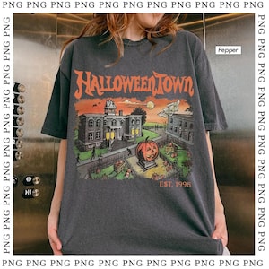 May include: Dark gray t-shirt with a Halloween-themed graphic. The design features a pumpkin, buildings, and the text "HALLOWEEN TOWN EST. 1998" in orange and white. The shirt has short sleeves.