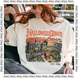 May include: Ivory-colored t-shirt with a Halloween-themed graphic. The design features the words "Halloween Town" in orange, along with a spooky town scene, a jack-o'-lantern, and the text "EST. 1998".