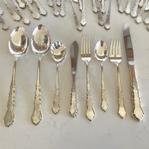 May include: A set of silver flatware, including spoons, forks, and a butter knife, with ornate handles. The flatware is arranged on a white surface.