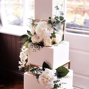 May include: A white square wedding cake with gold geometric tiers decorated with white flowers and greenery.