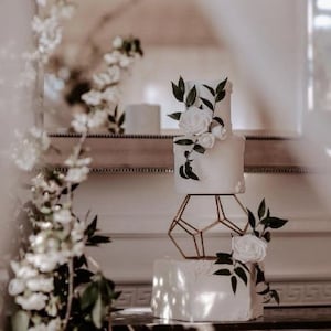May include: A white wedding cake with white flowers and green leaves on each tier. The cake is on a geometric gold stand.