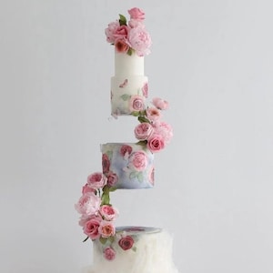 Puede incluir: Un pastel de boda blanco de cuatro pisos decorado con flores de azúcar rosas y blancas. El piso inferior es un pastel con volantes con glaseado blanco. Los tres pisos superiores están decorados con patrones florales y flores de azúcar rosas y blancas.