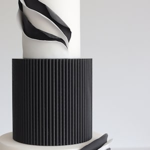 May include: A three-tiered wedding cake with black and white fondant. The top tier has a white fondant base with black fondant waves. The middle tier is black fondant with vertical lines. The bottom tier is white fondant with black fondant waves.
