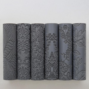 Puede incluir: Seis cilindros grises con patrones en relieve. Los diseños incluyen un patrón de hojas, encaje adornado y motivos florales. Los cilindros están dispuestos verticalmente sobre un fondo blanco, mostrando las texturas detalladas.