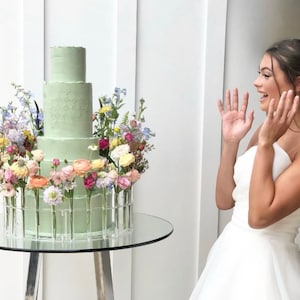 May include: A three-tiered light green wedding cake decorated with fresh flowers in a variety of colors, including pink, yellow, blue, and purple. The cake is displayed on a clear glass cake stand.