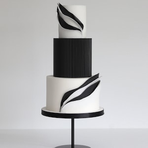 May include: A three-tier wedding cake decorated with black and white fondant. The bottom tier is white with black fondant waves, the middle tier is black with vertical lines, and the top tier is white with black fondant waves. The cake is on a black cake stand.