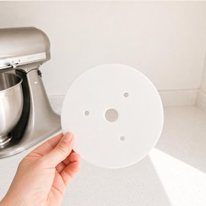 May include: A circular white flat disc featuring a large center hole and three smaller holes arranged in a triangular pattern around the middle. The object is made of a smooth, opaque material and has a clean, simple design for functional use.